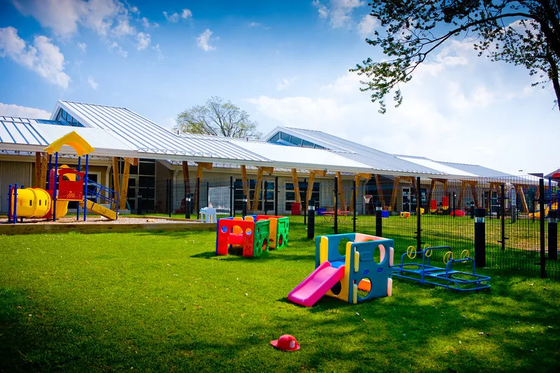 Colorful outdoor play area at a childcare center, with slides, climbing equipment and toy vehicles on a green lawn beside a yellow childcare building under a blue sky