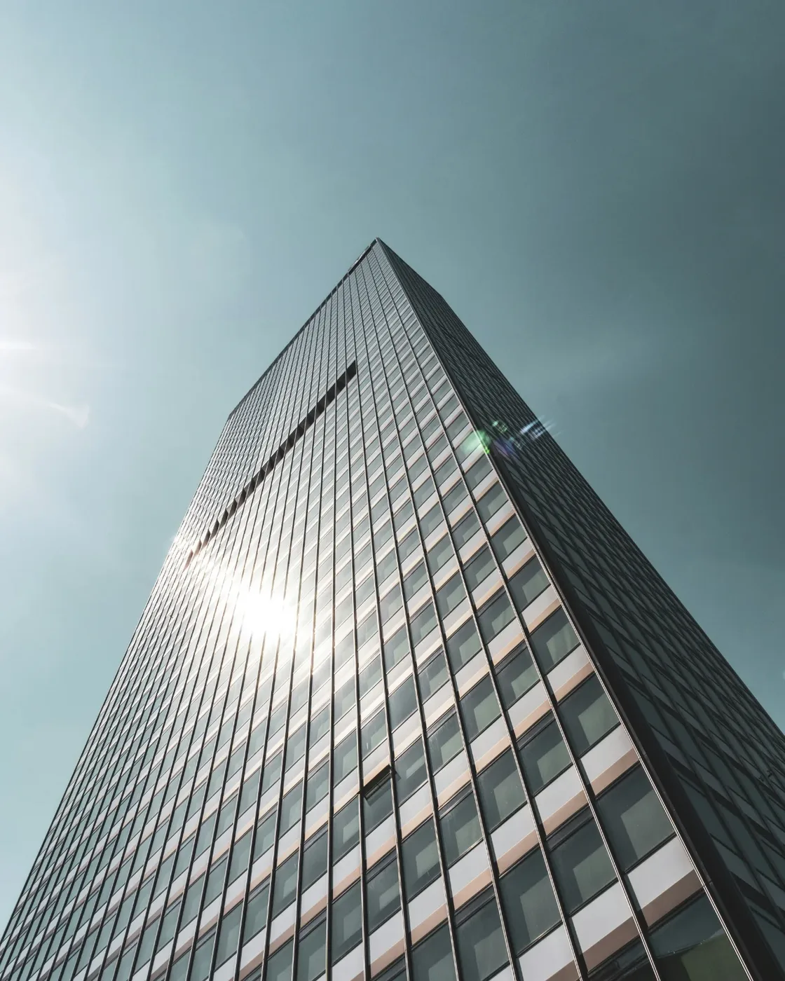 Modern glass and steel office tower seen from below against a clear, hazy sky, sunlight flaring off the glass